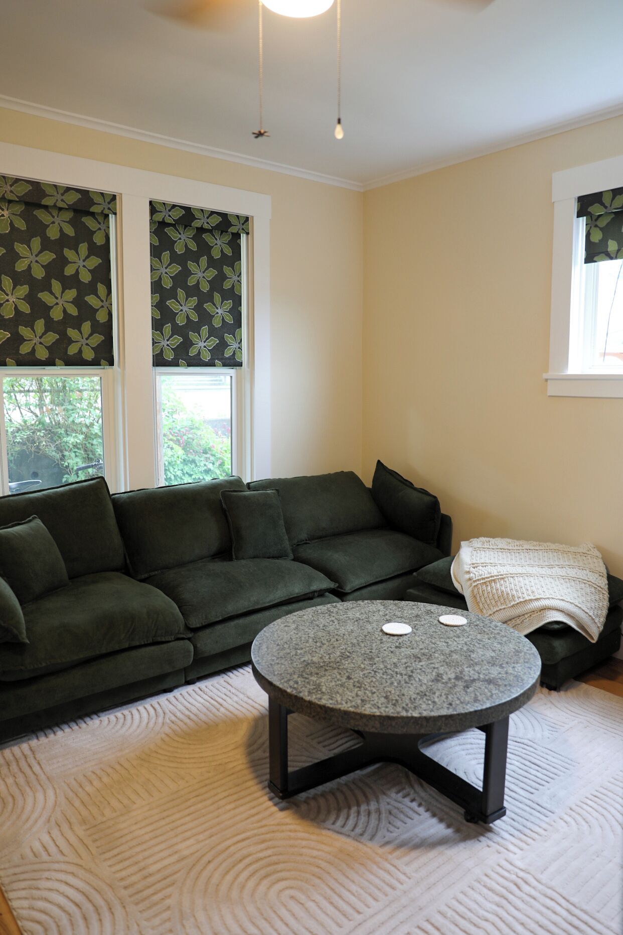 A living room with a sectional sofa, a round coffee table, and a window with a view of greenery.