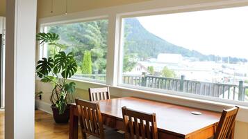 A dining area with a wooden table and chairs, a ceiling fan, and a view of greenery and buildings through large windows.