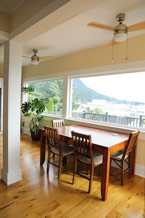A dining area with a wooden table and chairs, a ceiling fan, and a view of greenery and buildings through large windows.