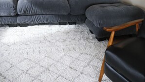 Un salon avec un canapé d’angle gris, une petite table en bois, un tableau représentant un paysage encadré et une lampe fixée au mur.