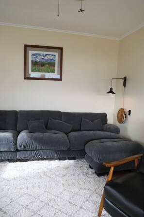 A living room with a grey sectional sofa, a small wooden table, a framed landscape painting, and a wall-mounted lamp.