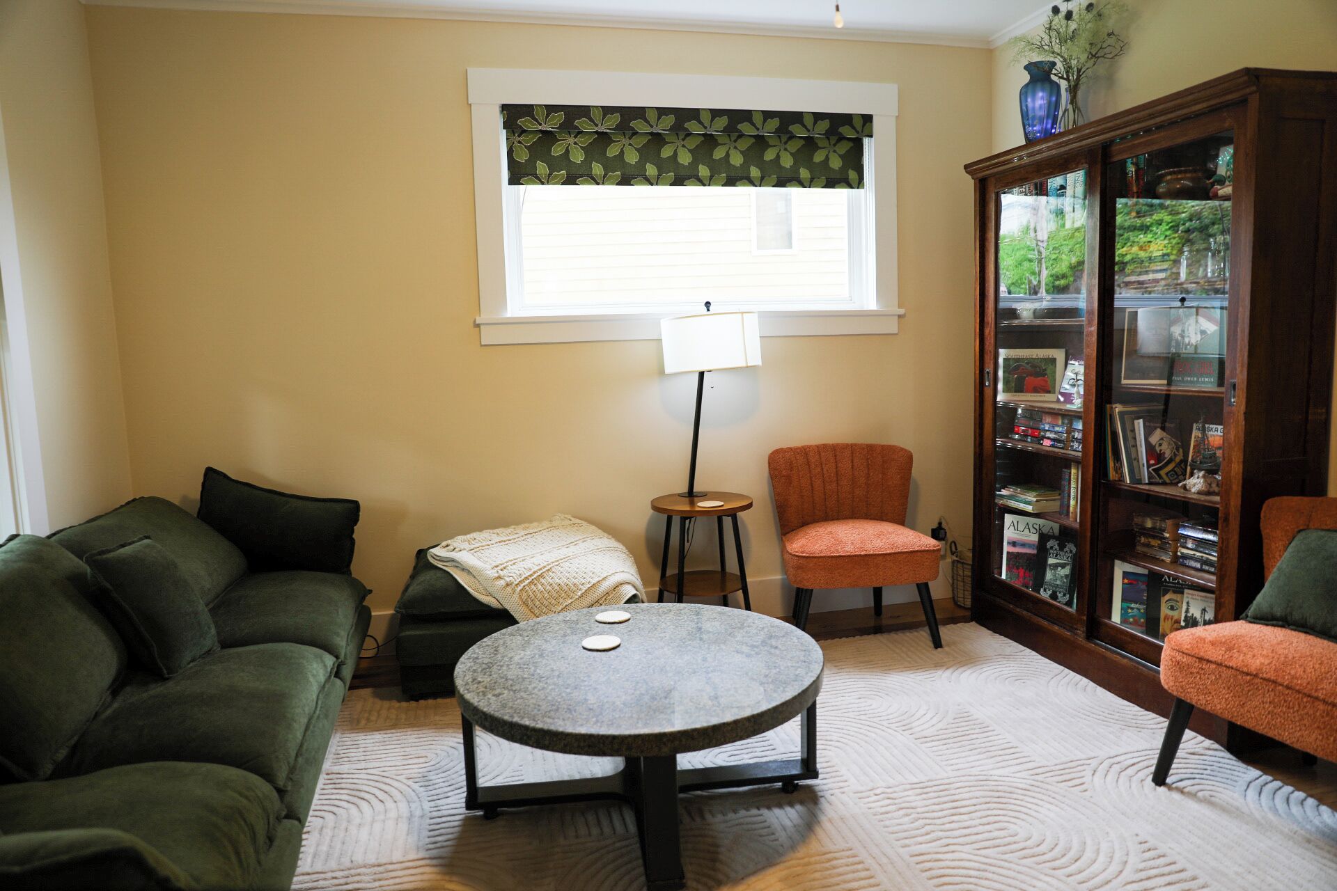 A living room with a green sofa, a round coffee table, a small side table with a lamp, a red armchair, and a bookshelf filled with books.