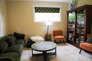 A living room with a green sofa, a round coffee table, a small side table with a lamp, a red armchair, and a bookshelf filled with books.