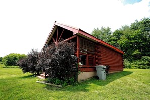Eine rote Holzhütte mit geneigtem Dach, umgeben von üppigem Grün und Bäumen.