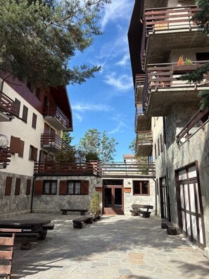 A courtyard with stone flooring, wooden benches, and a building with balconies and windows. - Mountain atmosphere at the ski lift departure area (Sauze d'Oulx)