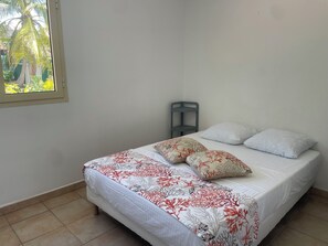 Ein Bett mit floralem Überwurf und zwei Kissen, ein Fenster mit Blick ins Grüne und ein Stuhl an der Wand.