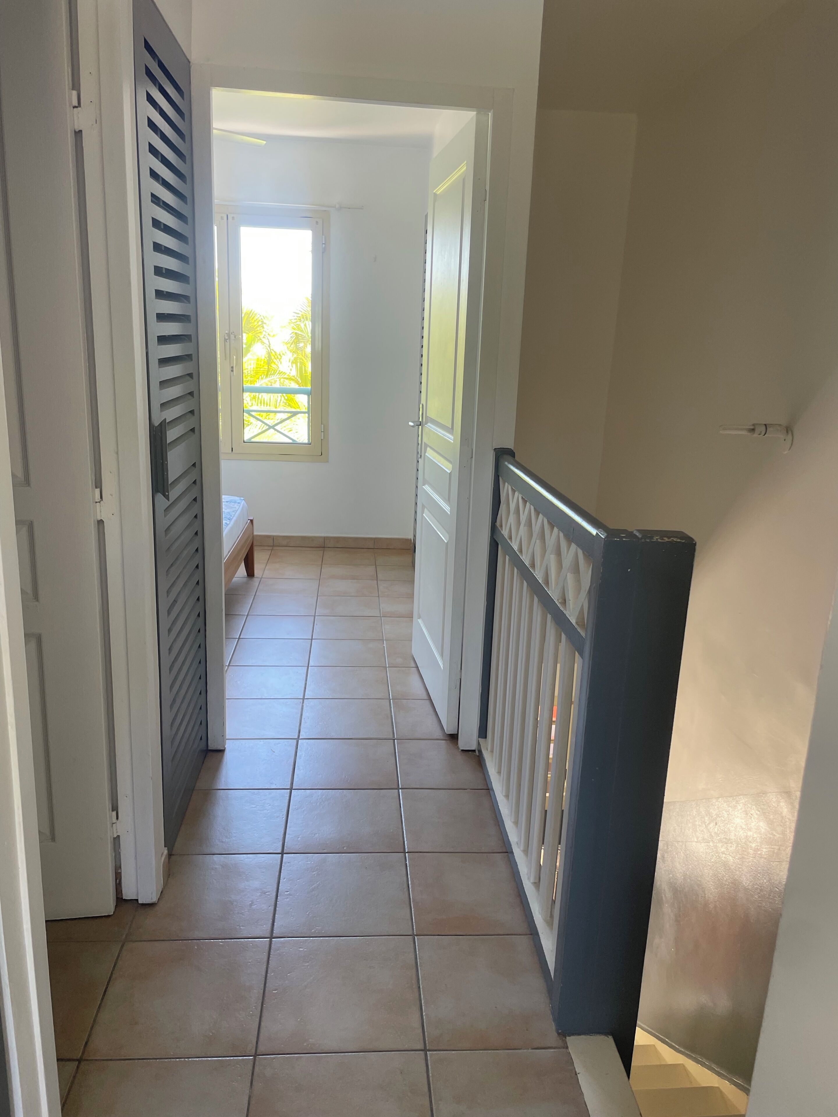 Un couloir au sol carrelé, une fenêtre donnant sur un espace verdoyant et une porte menant à un balcon.