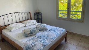 Ein Bett mit Holz Kopfteil, weißer Bettwäsche und blau gepolsterten Kissen mit Blumenmuster. Ein kleiner Tisch mit einer Lampe steht neben dem Bett.