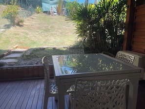Een terras buiten met een tafel met glazen blad en witte stoelen, omringd door een houten terras en een gazon met planten.