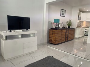 Ein modernes Wohnzimmer mit Flachbildfernseher, einer weißen Unterhaltungsecke, einer Küchenzeile mit weißen Schränken und einem gefliesten Boden.