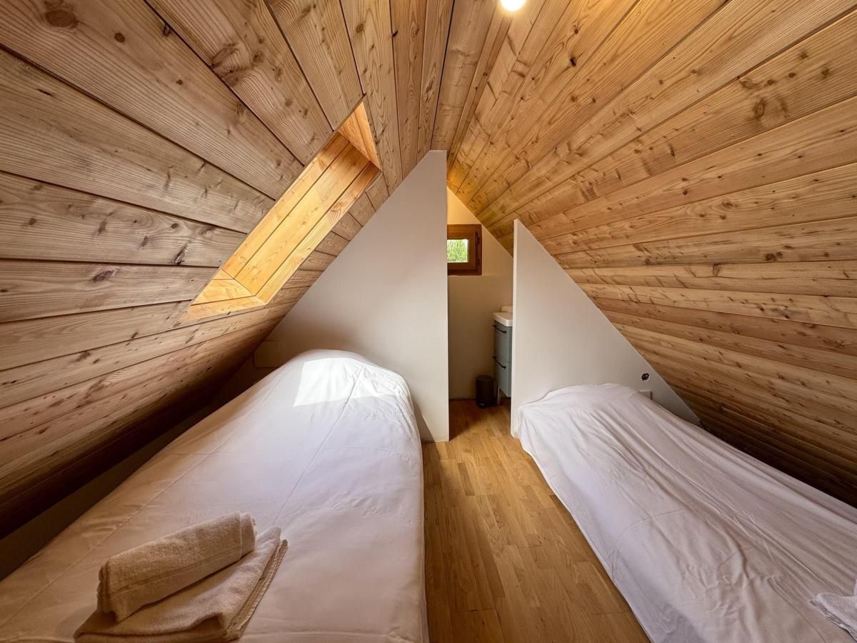 Une petite chambre mansardée, chaleureuse et accueillante, avec des murs en bois et un plafond en pente. On y trouve deux lits simples avec du linge de lit blanc et une petite fenêtre qui laisse entrer la lumière naturelle.