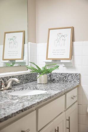 Un baño con encimera de granito, un lavabo, un espejo y dos cuadros en la pared.
