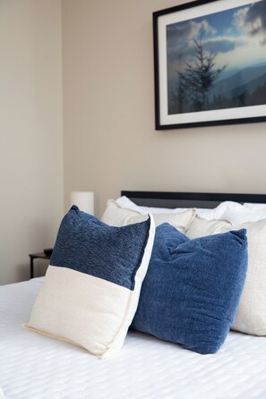 Una cama con almohadas blancas y azules, un cuadro enmarcado en la pared y una mesita de noche con una lámpara.