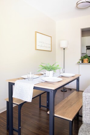 Un comedor con una mesa de madera preparada para dos personas, una planta en maceta y una cita enmarcada en la pared.