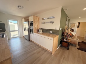 A modern kitchen with a built-in refrigerator, oven, and coffee maker. There is a dining area with a chair and a decorative plant. - GatherRound | Groups, Family, Spa | Flexible Beds | SQ PROPERTY (Palmerston North)
