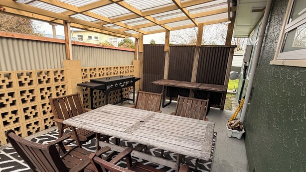 An outdoor patio with a wooden table and chairs, a grill, and a patterned rug. - SQ Property; Autumn Hideaway. Customisable layout for small or large groups (Palmerston North)