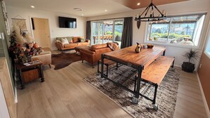 A modern living room with a wooden dining table, leather sofas, and a large window with a view. - SQ Property; Autumn Hideaway. Customisable layout for small or large groups (Palmerston North)