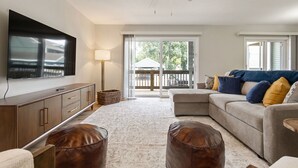 A living room with a flat-screen TV, a ceiling fan, a sofa with cushions, a coffee table, and a large window with a view of the outdoors.