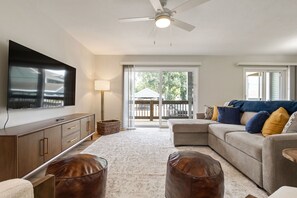 Una sala de estar con televisor de pantalla plana, ventilador de techo, sofá con cojines, una mesa de centro y un ventanal con vistas al exterior.