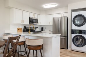 A modern kitchen with white cabinetry, stainless steel appliances, a central island with bar stools, and a built-in washer and dryer. - Stylish ND Haven | 2B/2BA | Balcony + Walk to ND (South Bend)
