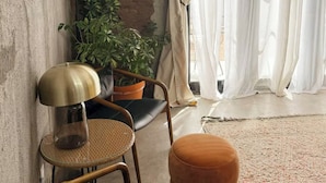 A room with a concrete wall, a potted plant, a chair, a small round table, and a large window with sheer curtains. - Bohemio Raconteur: Four Suites + Sauna + Pickleball + Walkable Marfa (Marfa)