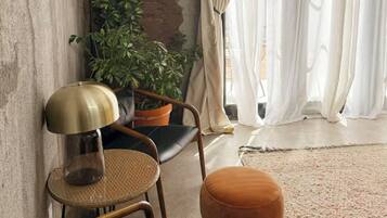 Une pièce avec un mur en béton, une plante en pot, une chaise, une petite table ronde et une grande fenêtre ornée de rideaux légers.