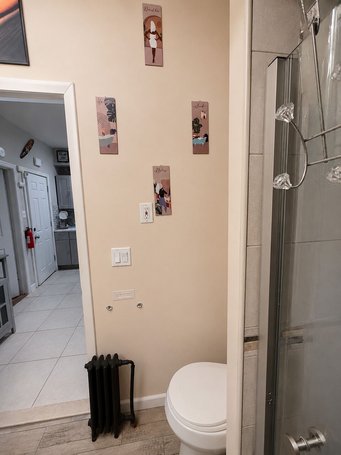 A bathroom with a toilet, a shower, and a radiator. There are wall-mounted light switches and a power outlet.