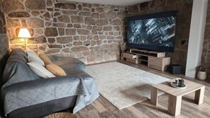 A living room with a stone wall, a sofa, a TV, and a coffee table. - Stone house with an olive grove, a movie theater, and a sim racing setup—sleeps 8 near Serra da Estrela (Distrito da Guarda)
