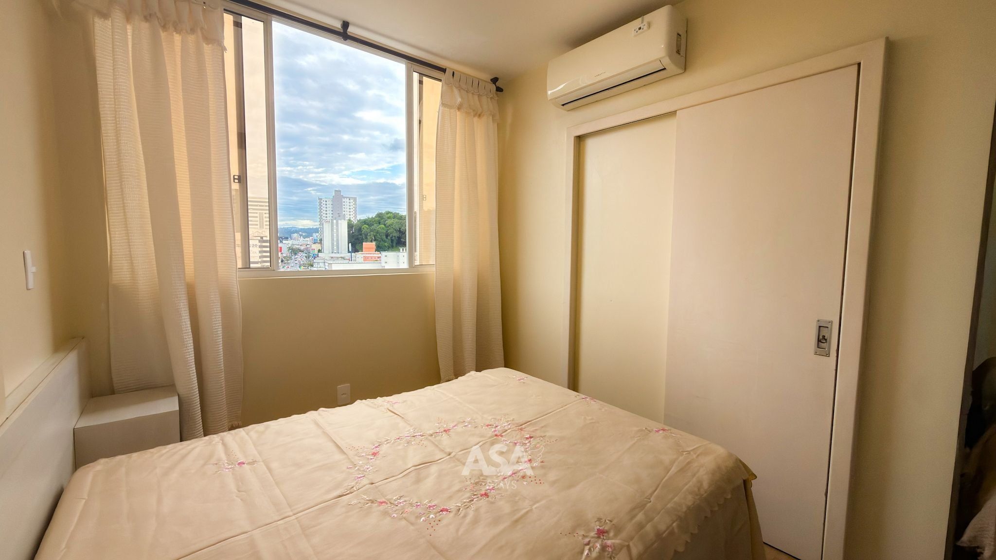 A bedroom with a bed, a window with curtains, an air conditioning unit, and a view of buildings outside.
