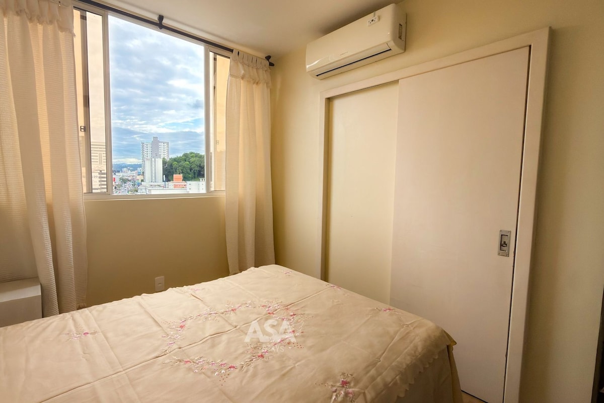 A bedroom with a bed, a window with curtains, an air conditioning unit, and a view of buildings outside.