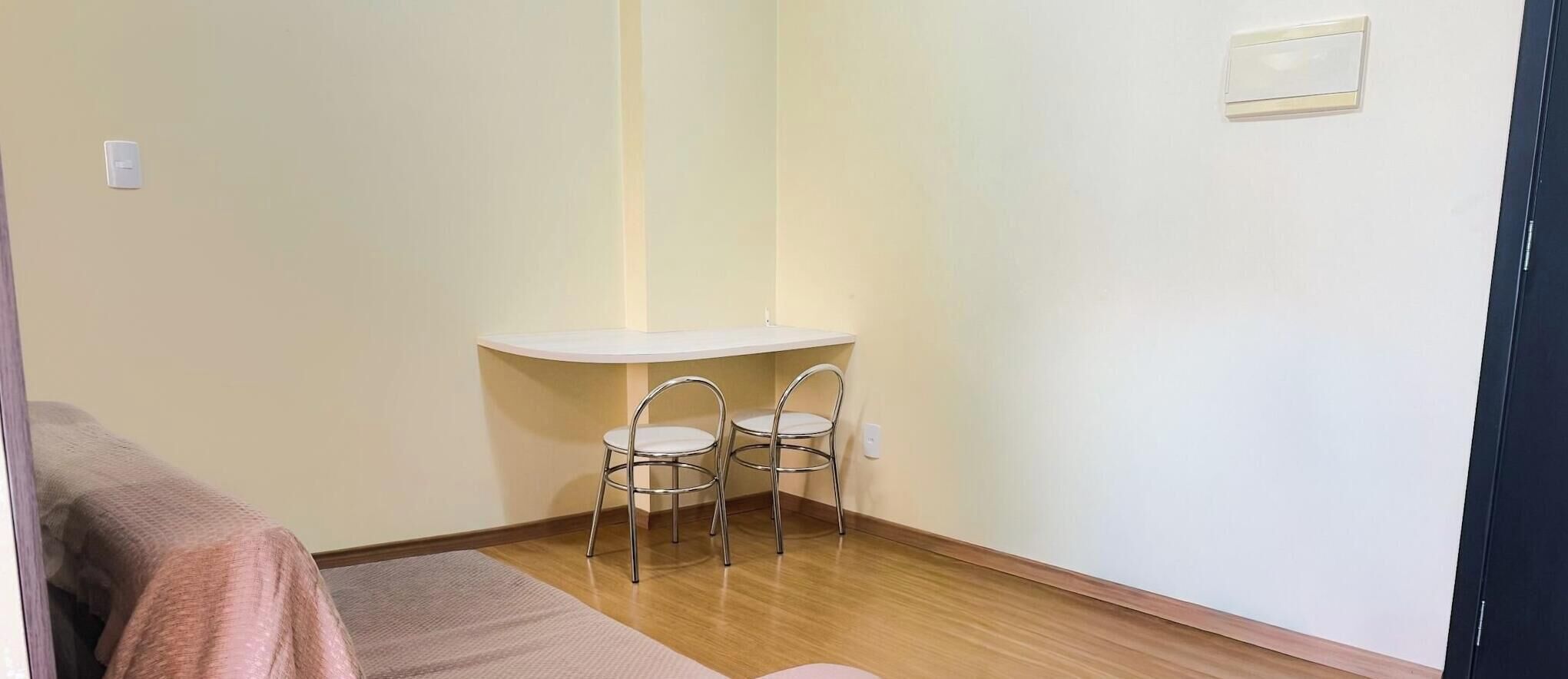 A small room with a wooden floor, a white wall, a white table with two chairs, and a bed with a pink blanket.