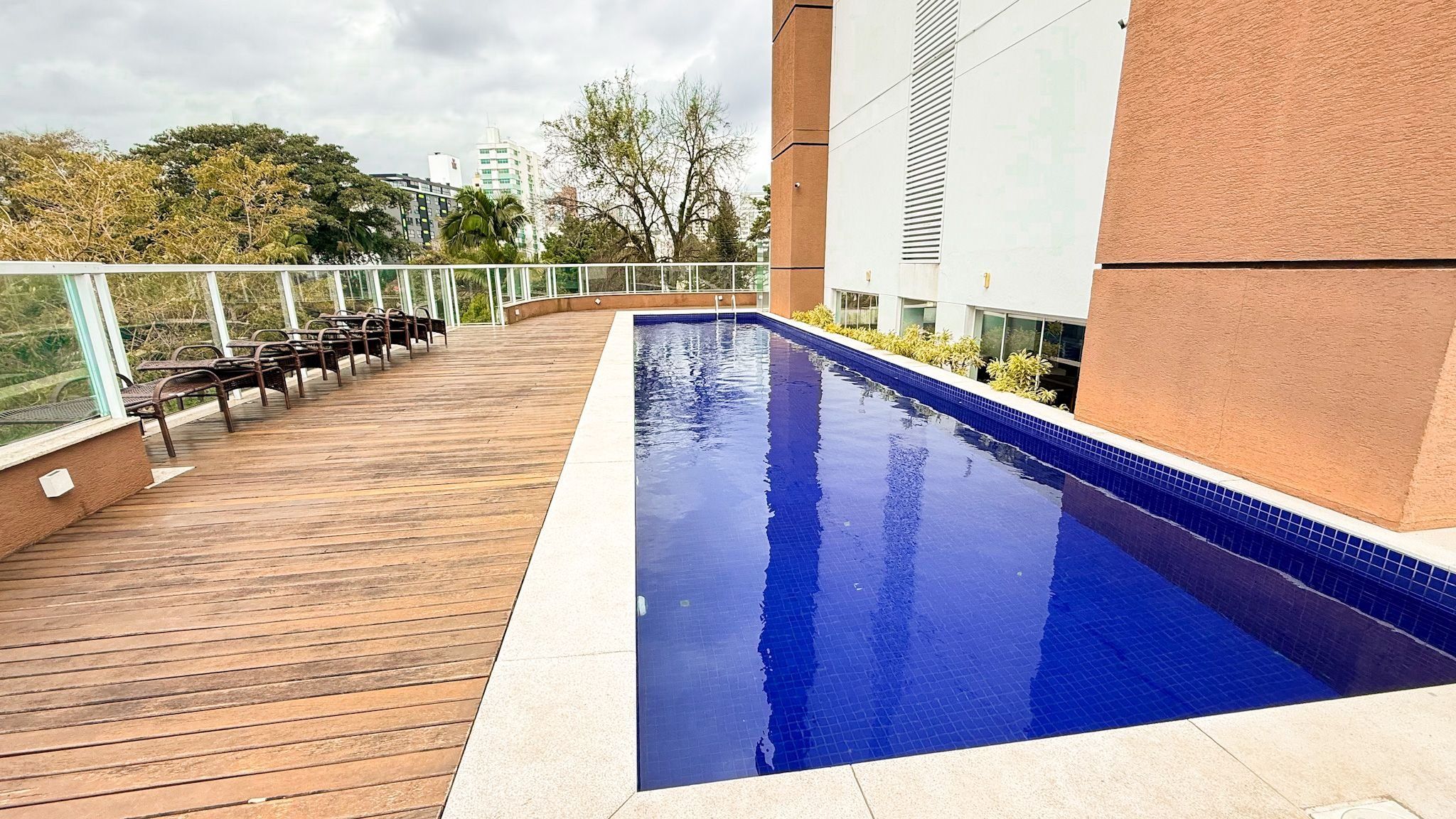 A swimming pool with a wooden deck and metal chairs, surrounded by a glass railing.