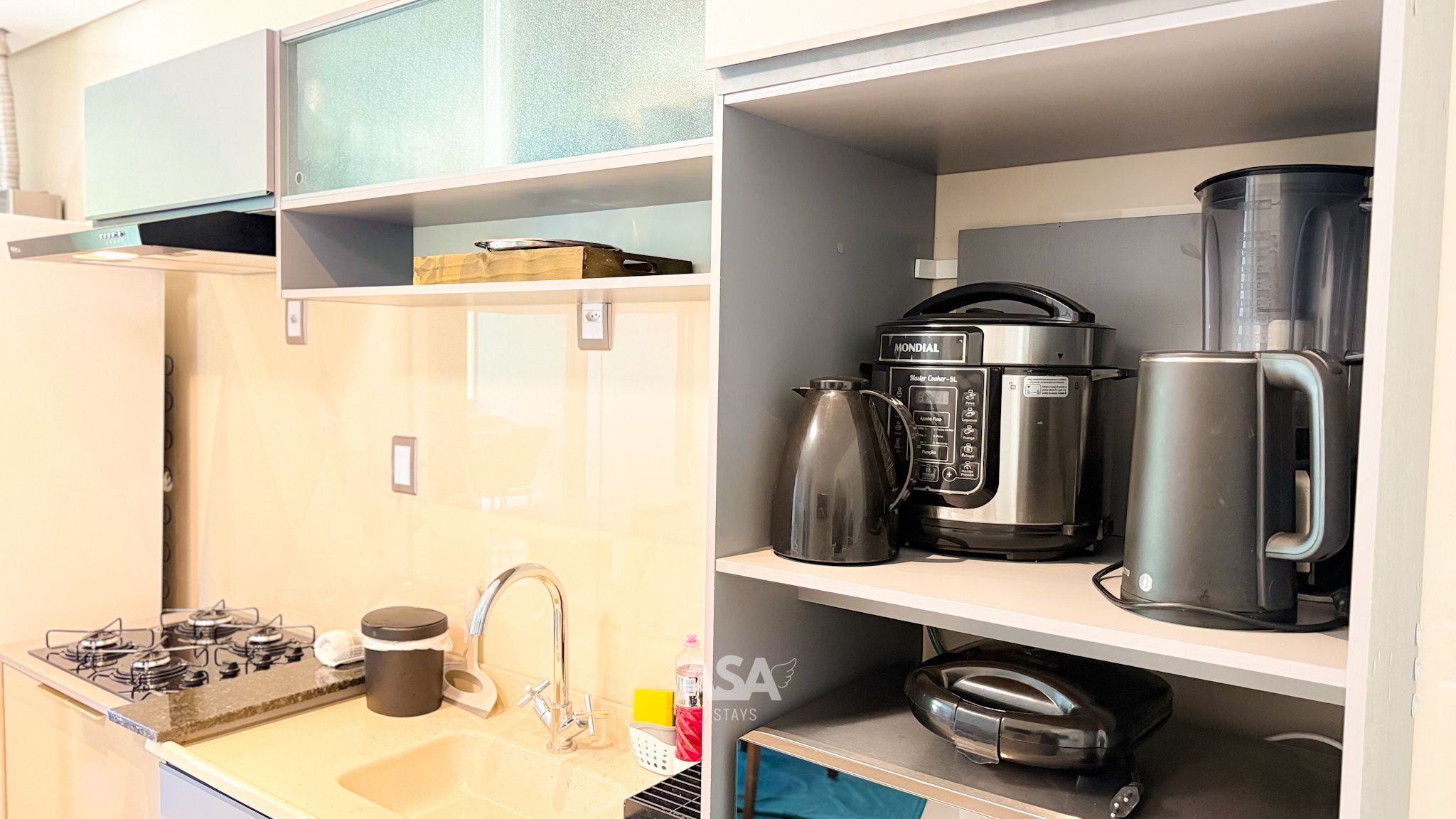 Une cuisine compacte équipée d’une cuisinière à gaz, d’une machine à café, d’une bouilloire et d’un mixeur, le tout rangé sur des étagères.
