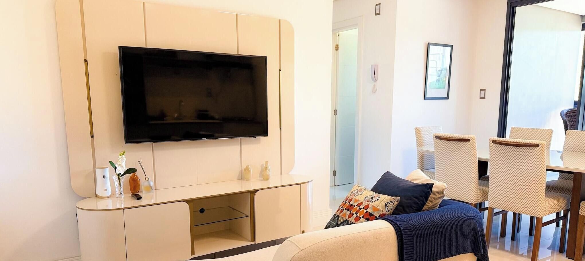 A living room with a flat-screen TV mounted on the wall, a sofa with cushions, a coffee table, and a dining area with chairs and a table.