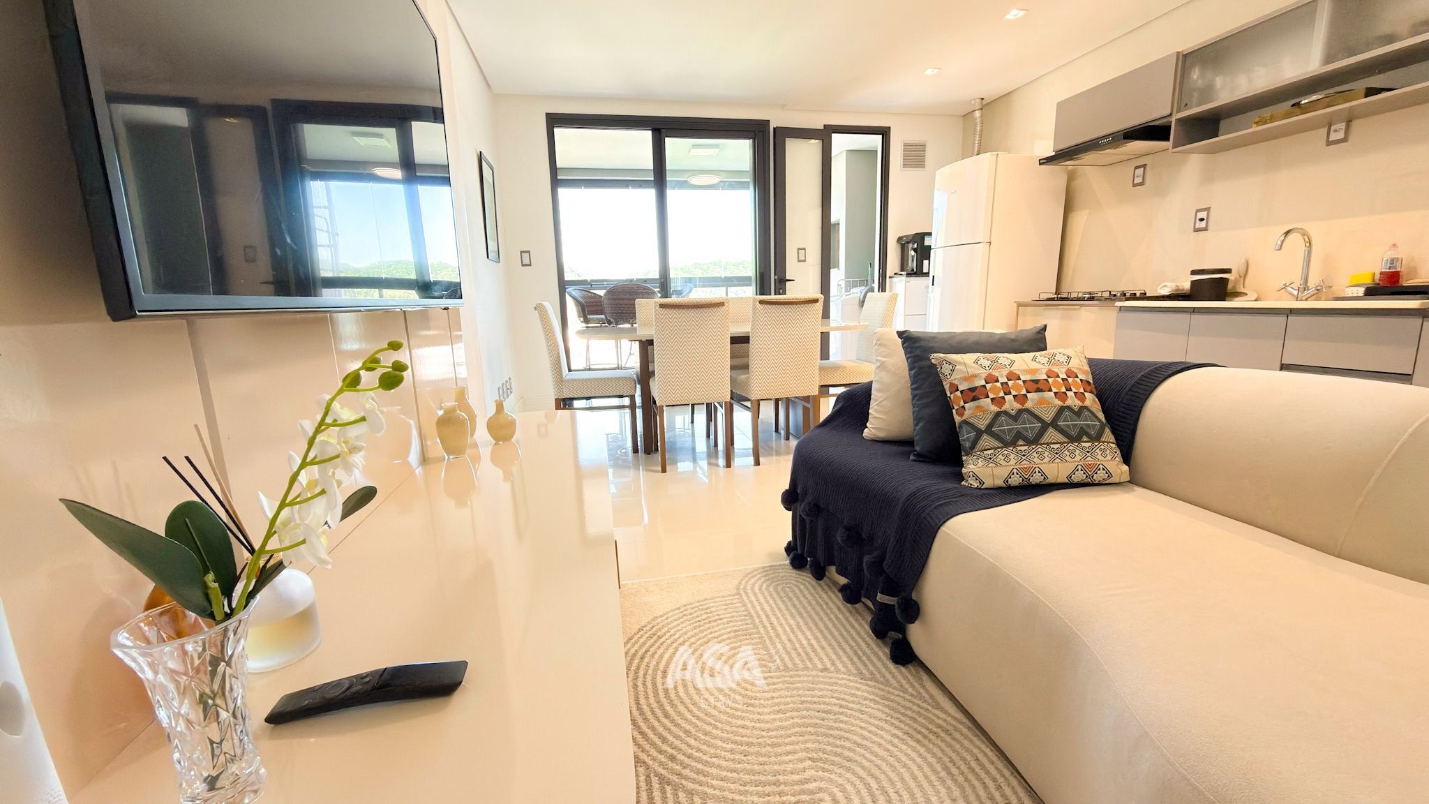 A modern living room with a sofa, a coffee table, and a TV mounted on the wall. There is a dining area with chairs and a table in the background.