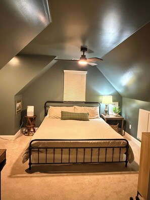 Un dormitorio con techo inclinado, una cama con cabecera, una mesita de noche con lámpara y una ventana con persianas.