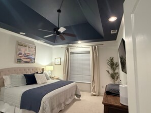 Un dormitorio con cama, ventilador de techo, mesita de noche, lámpara, ventana con persianas y una planta.