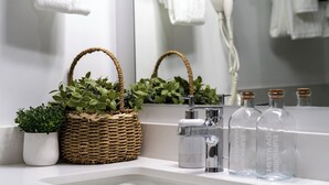 Ett badrum med ett vitt handfat, en spegel, en flätad korg med gröna växter, en modern blandare och genomskinliga glasflaskor med korkproppar.