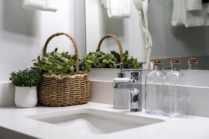 A bathroom with a white sink, a mirror, a woven basket with green plants, a modern faucet, and clear glass bottles with cork stoppers. - SOL Euphoria Ocho Rios walk to the beach (Ocho Rios)