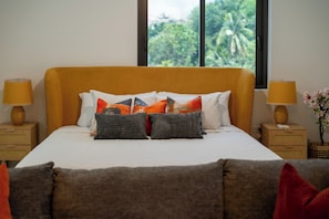 A bed with a headboard, multiple pillows, two bedside lamps, and a view of greenery through the window. - SOL Euphoria Ocho Rios walk to the beach (Ocho Rios)
