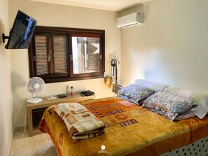 Quarto com cama, ventilador, televisão, janela com persianas, ar condicionado e mesa de cabeceira com abajur e chapéu.