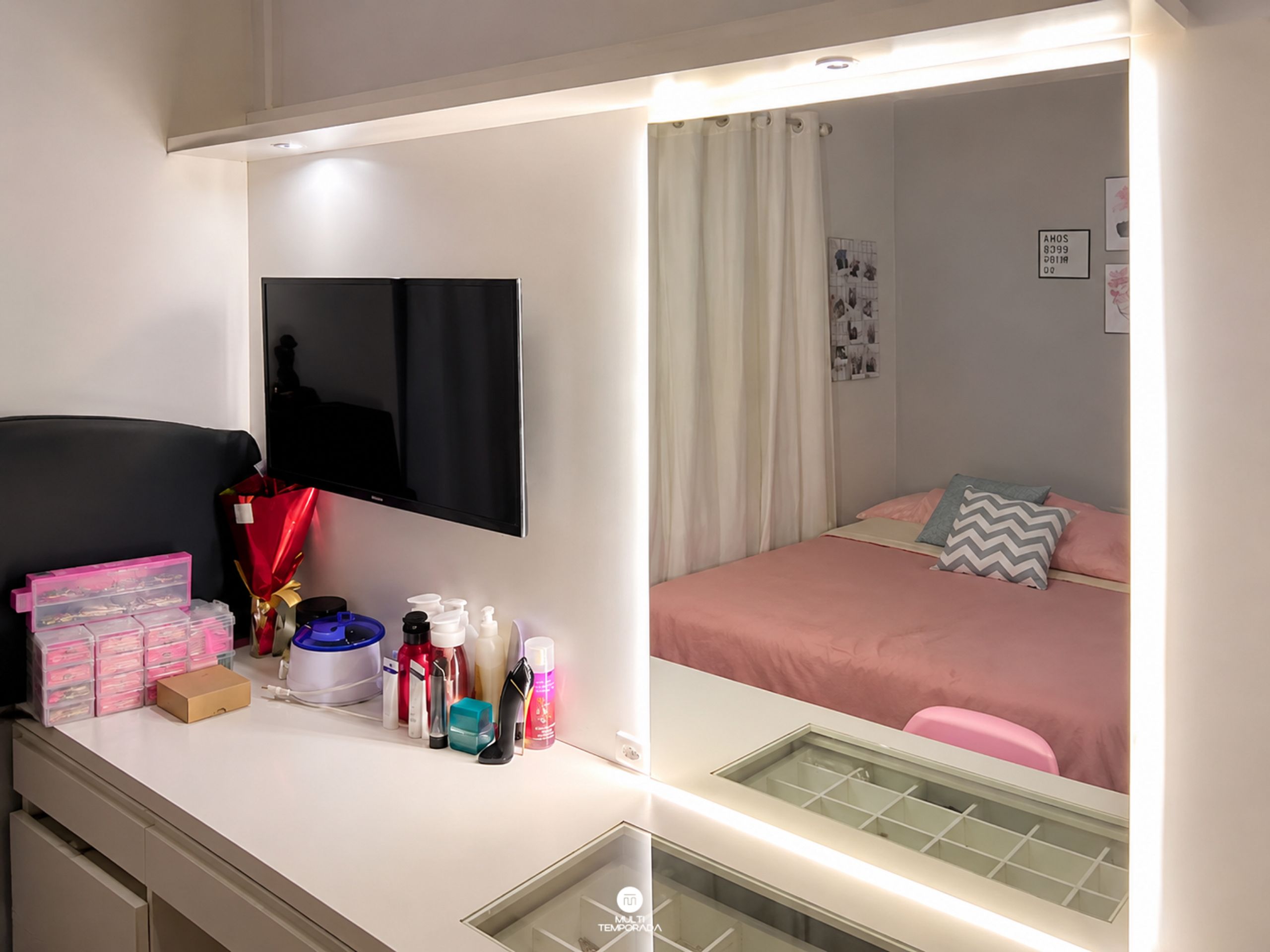 Quarto com cama, televisão, escrivaninha e vários objetos, e um espelho.