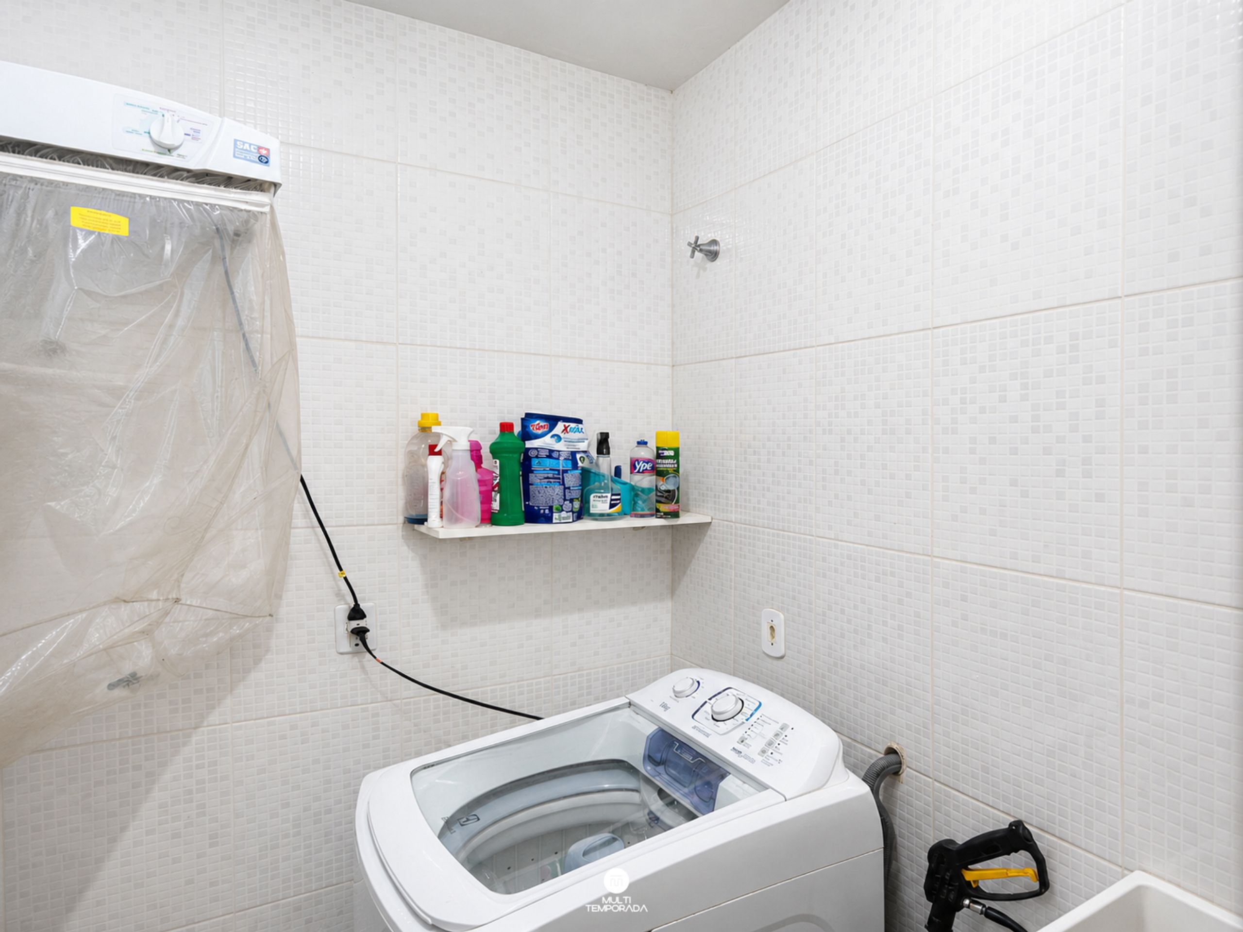 Uma lavanderia com máquina de lavar, prateleira com produtos de limpeza e área de chuveiro.
