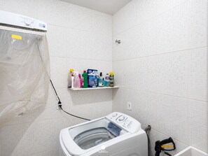 Uma lavanderia com máquina de lavar, prateleira com produtos de limpeza e área de chuveiro.