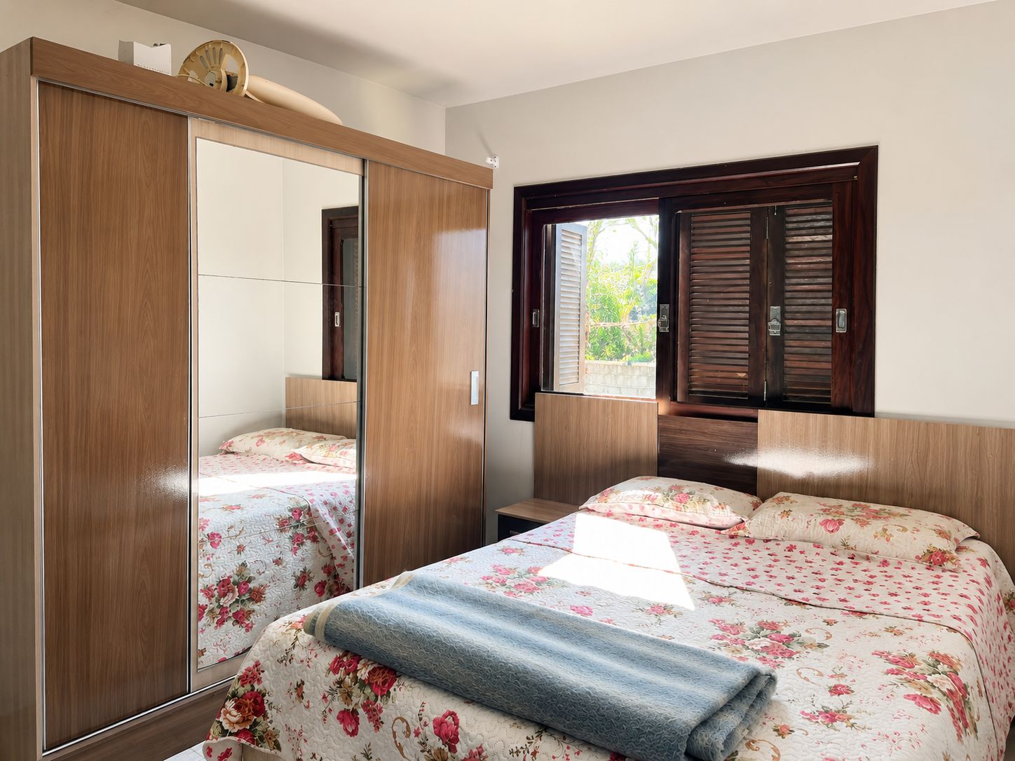 A bedroom with a wooden wardrobe, a bed with floral bedding, and a window with shutters.
