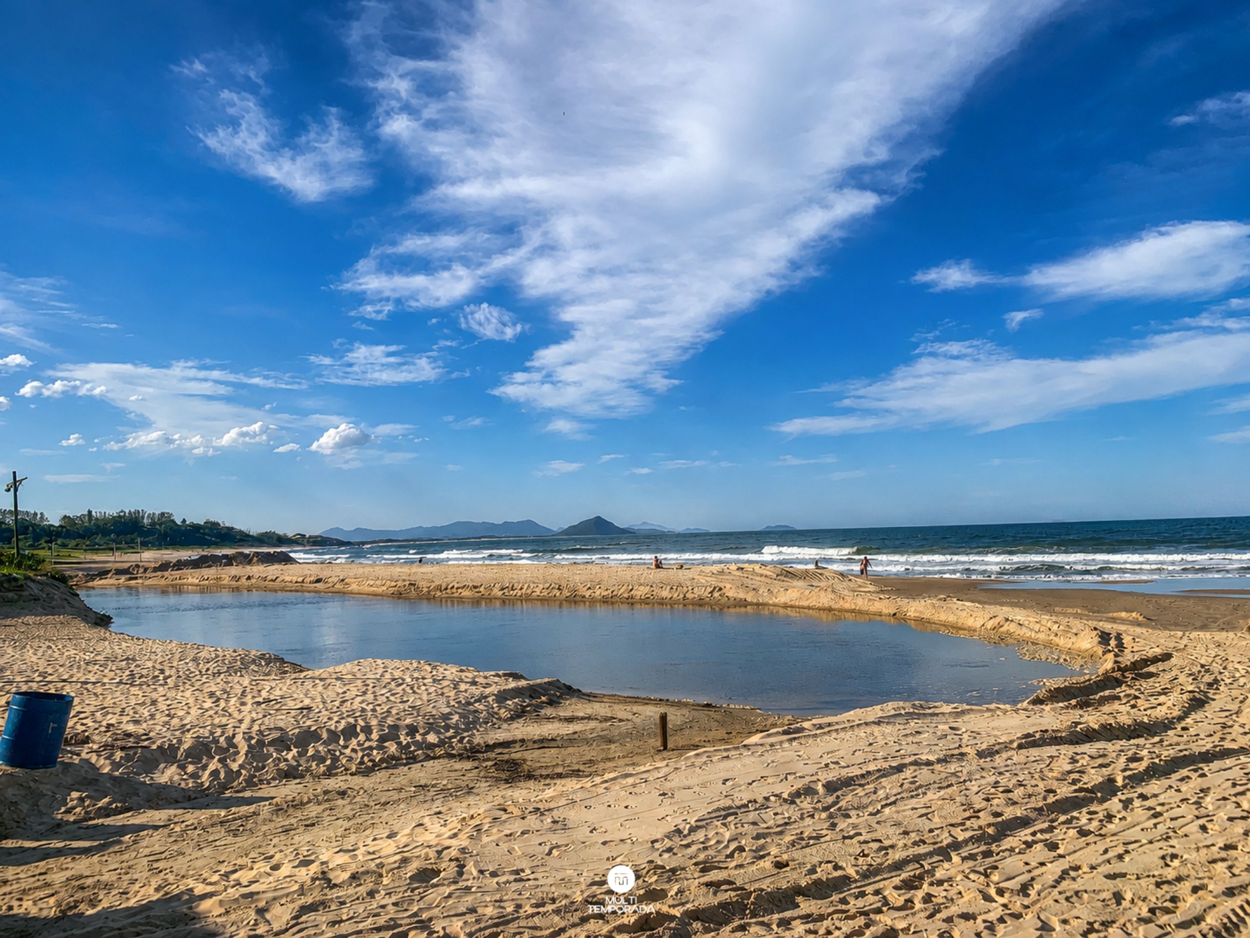 Uma praia com um pequeno lago, terreno arenoso e montanhas ao longe.