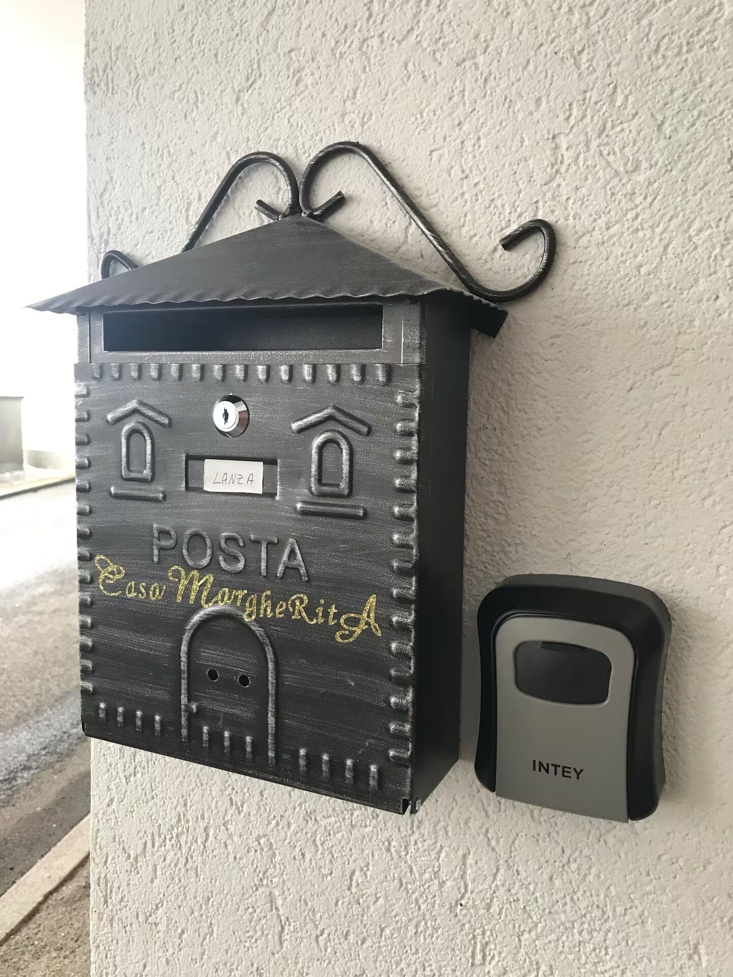 A mailbox with a house-like design, labeled 'POSTA Caso Margherita,' mounted on a wall next to a door with an 'INTEY' label.