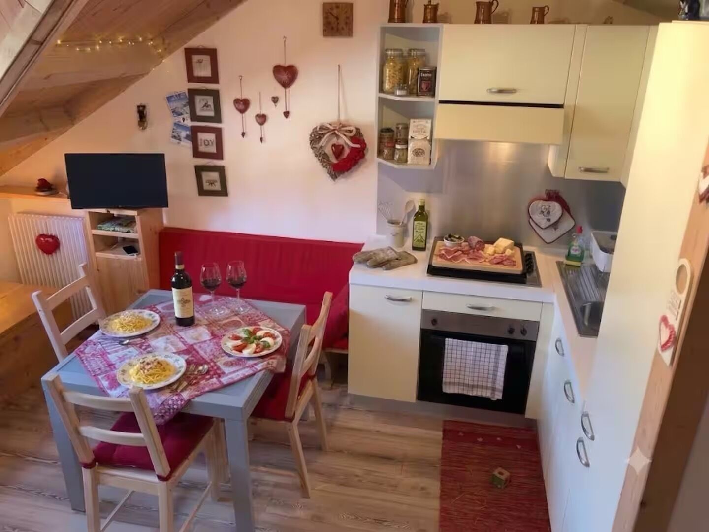 Un'accogliente zona pranzo con un tavolo apparecchiato per due, una panca imbottita rossa e un angolo cottura con frigorifero, forno e diversi alimenti.