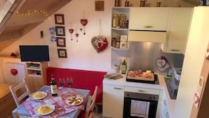 Un coin repas accueillant avec une table dressée pour deux personnes, un banc rembourré de couleur rouge et un espace cuisine équipé d’un réfrigérateur, d’un four et de divers produits alimentaires.