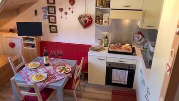 Un'accogliente zona pranzo con un tavolo apparecchiato per due, una panca imbottita rossa e un angolo cottura con frigorifero, forno e diversi alimenti.